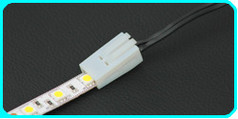 Wei�er LED-Verbinder mit Klemme, der eine LED-Leiste mit schwarzen Kabeln verbindet.