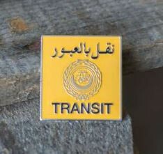 Quadratischer gelber Scandi Pin mit schwarzem "TRANSIT" und arabischem Schriftzug sowie silbernem Emblem.