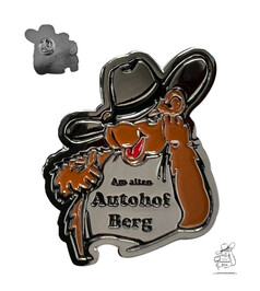 Metall-Anstecknadel in Form eines braunen Cartoon-B�ren mit grauem Shirt und der Aufschrift "Am alten Autohof Berg".